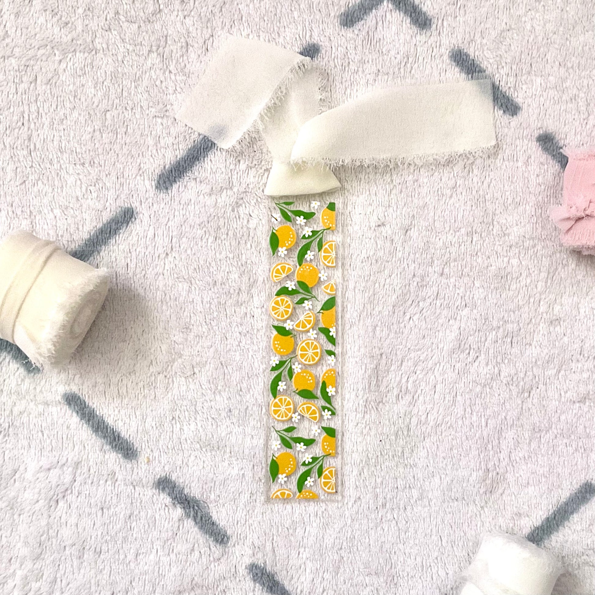 Clear bookmark made of acrylic, we can see through, with small full oranges and quarters of oranges and small white flowers and green orange tree leaves. Attached to it is a cream colour chiffon ribbon.