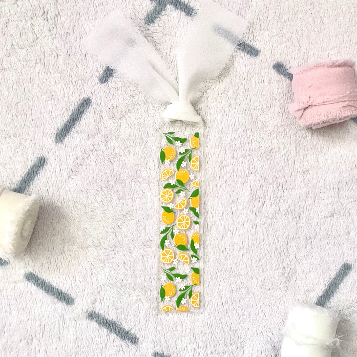 Clear bookmark made of acrylic, we can see through, with small full oranges and quarters of oranges and small white flowers and green orange tree leaves. Attached to it is a white colour chiffon ribbon.