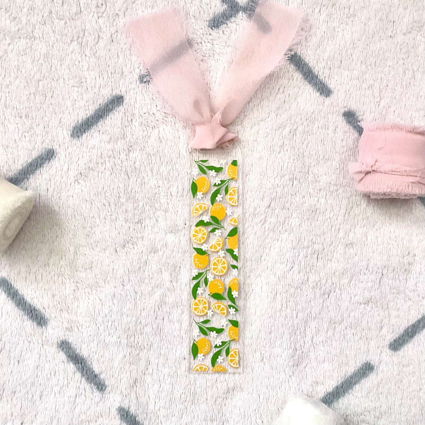 Clear bookmark made of acrylic, we can see through, with small full oranges and quarters of oranges and small white flowers and green orange tree leaves. Attached to it is a pale pink colour chiffon ribbon.