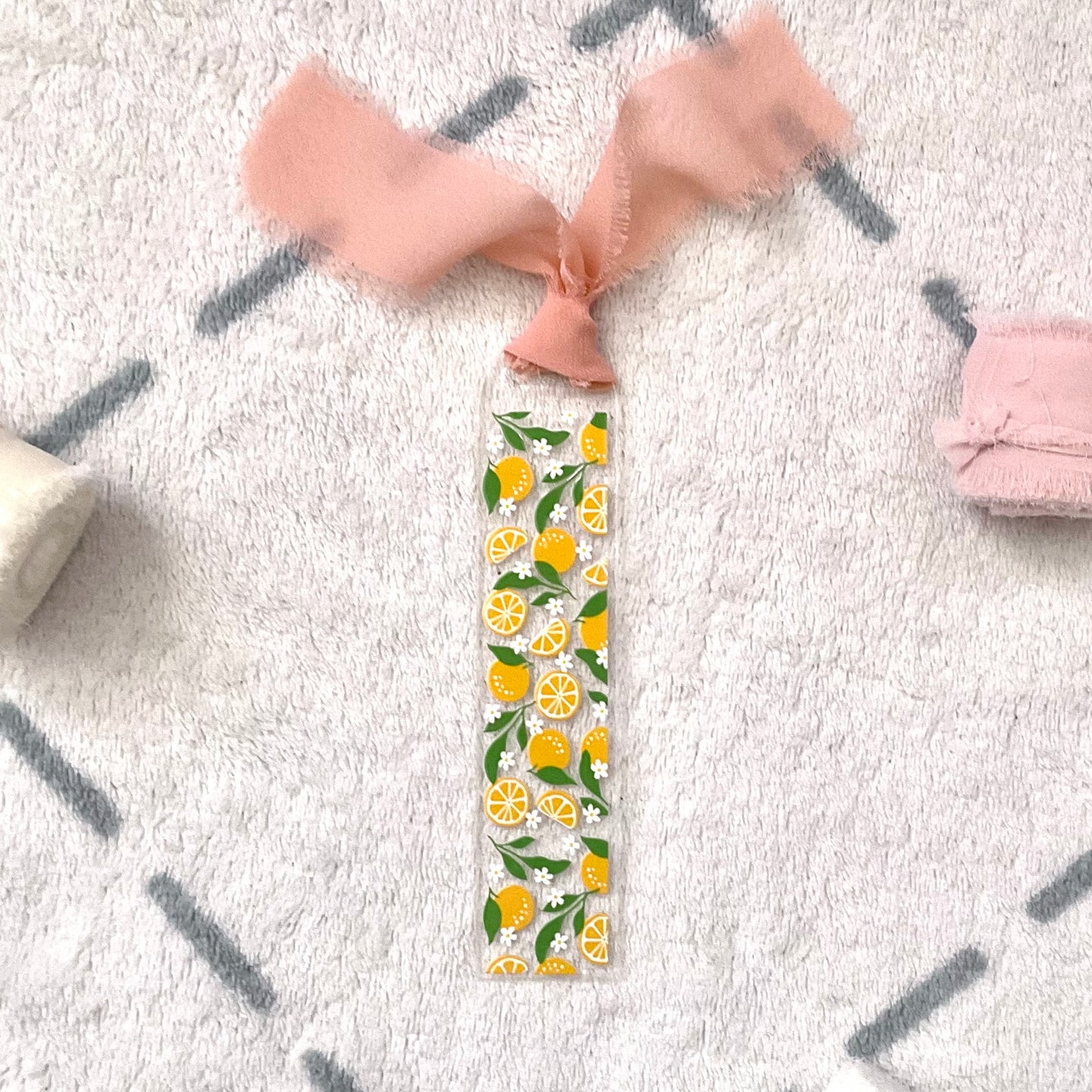 Clear bookmark made of acrylic, we can see through, with small full oranges and quarters of oranges and small white flowers and green orange tree leaves. Attached to it is a saumon (mix between pink and orange) colour chiffon ribbon.