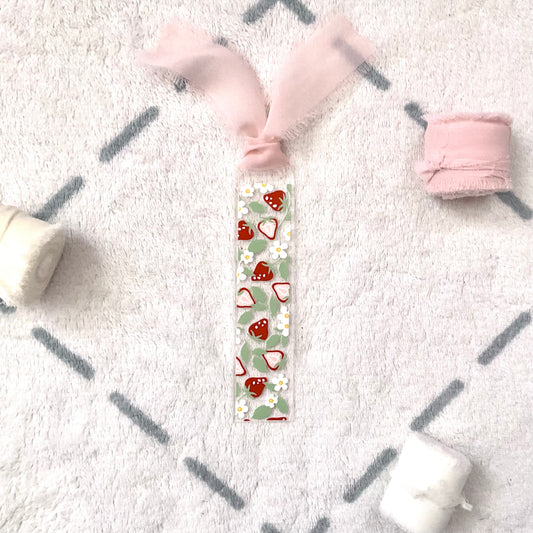 Clear bookmark made of acrylic, we can see through, with small red strawberries and small white flowers and pale green strawberry leaves. Attached to it is a pale pink chiffon ribbon. 