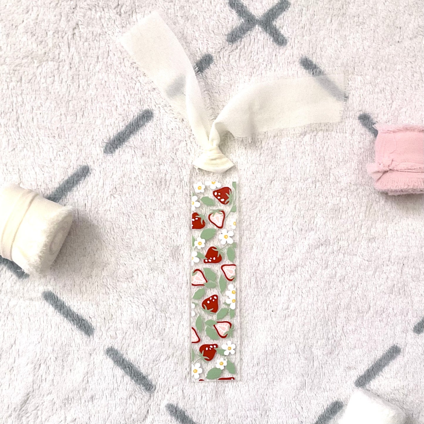 Clear bookmark made of acrylic, we can see through, with small red strawberries and small white flowers and pale green strawberry leaves. Attached to it is a cream chiffon ribbon.