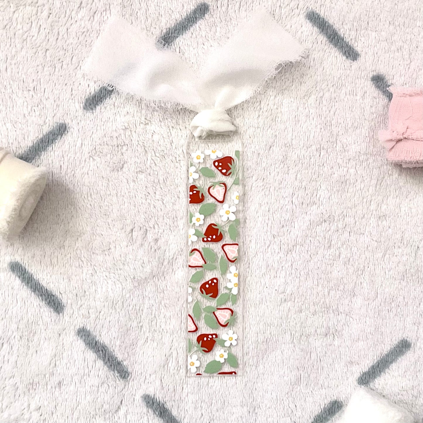 Clear bookmark made of acrylic, we can see through, with small red strawberries and small white flowers and pale green strawberry leaves. Attached to it is a white chiffon ribbon.