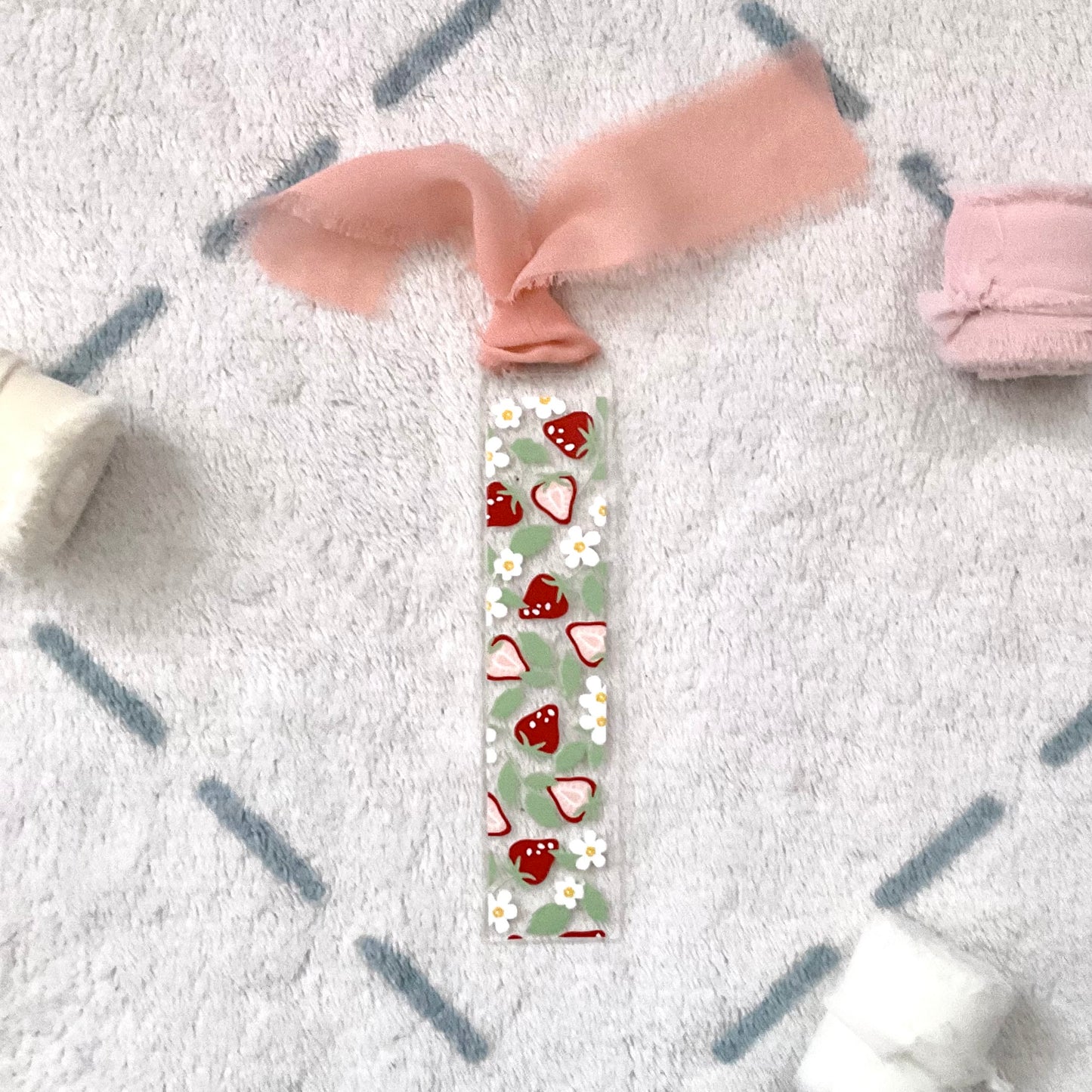 Clear bookmark made of acrylic, we can see through, with small red strawberries and small white flowers and pale green strawberry leaves. Attached to it is saumon colour (mix between pink and orange) chiffon ribbon.