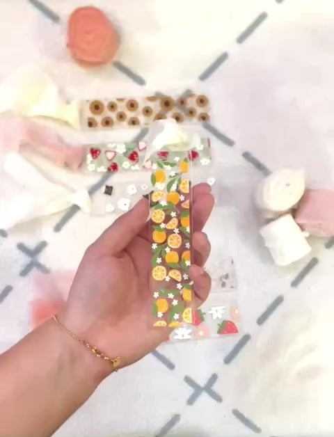 Short video that shows the bookmark made of acrylic. We can see through it, it has small full oranges and quarters of oranges and small white flowers and green orange tree leaves. Attached to it is a cream colour chiffon ribbon. When the bookmark is flipped to the other side the designs previously described are white shapes.