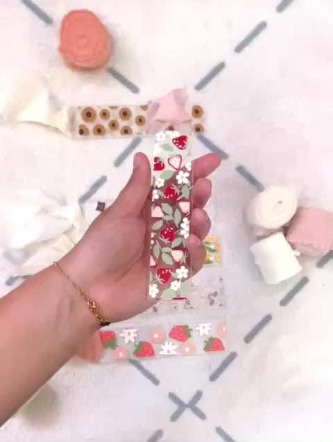 Short video that shows the bookmark made of acrylic. We can see through it, it has small red strawberries and small white flowers and pale green strawberry leaves designed on it. Attached to it is a pale pink chiffon ribbon. When the bookmark is flipped to the other side the designs previously described are white shapes.